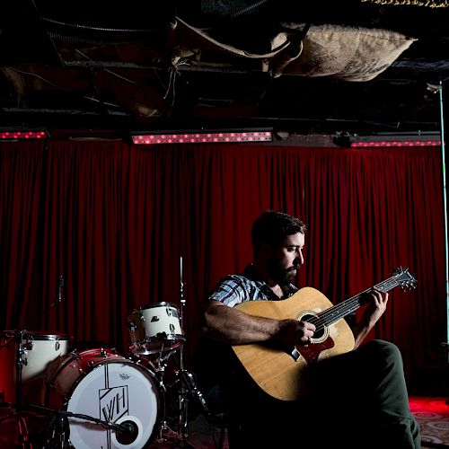 A man is playing an acoustic guitar in front of a red curtain with a drum set beside him.