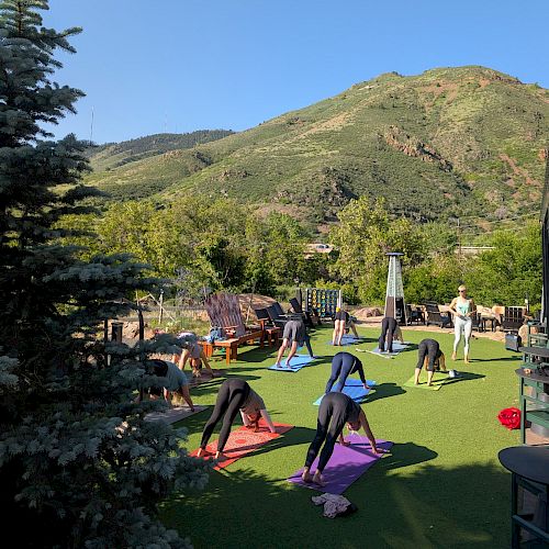 People are practicing yoga on colorful mats outdoors, surrounded by trees and mountains under a clear blue sky.