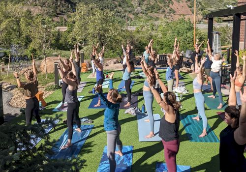 A group of people practicing yoga outdoors on mats, with a mountain in the background, under a clear blue sky.