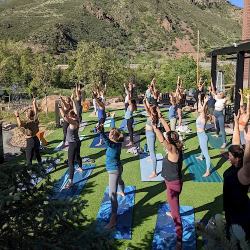 A group of people practicing yoga outdoors on mats, with a mountain in the background, under a clear blue sky.