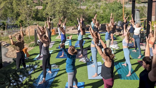 A group of people practicing yoga outdoors on mats, with a mountain in the background, under a clear blue sky.