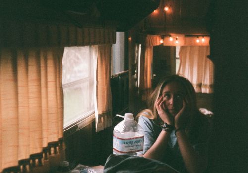 A person sits inside a warmly lit RV, with orange string lights and closed curtains, next to a large water jug and some personal items on a table.