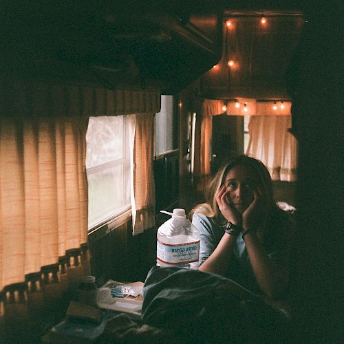 A person sits inside a warmly lit RV, with orange string lights and closed curtains, next to a large water jug and some personal items on a table.