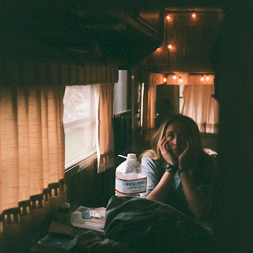 A person sits inside a warmly lit RV, with orange string lights and closed curtains, next to a large water jug and some personal items on a table.