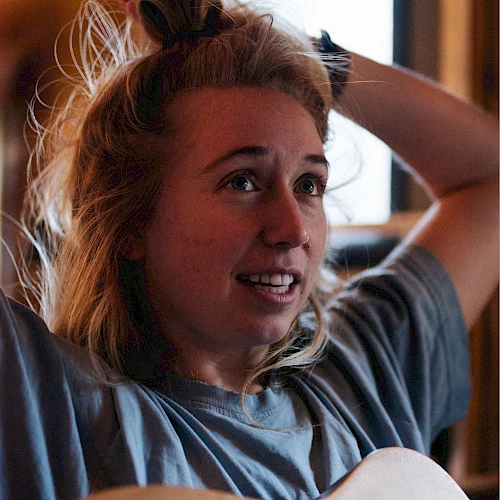 A person holds their hair up with one hand, smiling slightly, and has a guitar resting against them, indoors with casual attire.