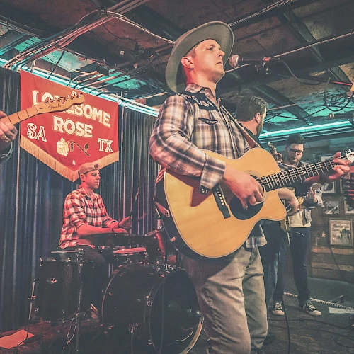 A band performs on stage, featuring a guitarist and drummer, in a venue with a banner reading "Lonesome Rose" in the background.