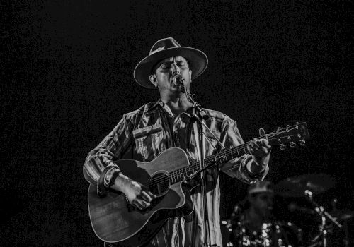 A musician wearing a hat plays an acoustic guitar and sings on stage, with a drummer in the background.