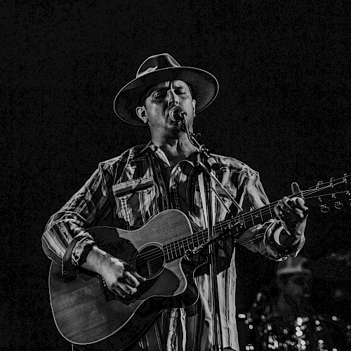 A musician wearing a hat plays an acoustic guitar and sings on stage, with a drummer in the background.