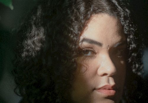 A person with curly hair is partially illuminated by light filtering through leaves, creating a dramatic, shadowed effect on their face.
