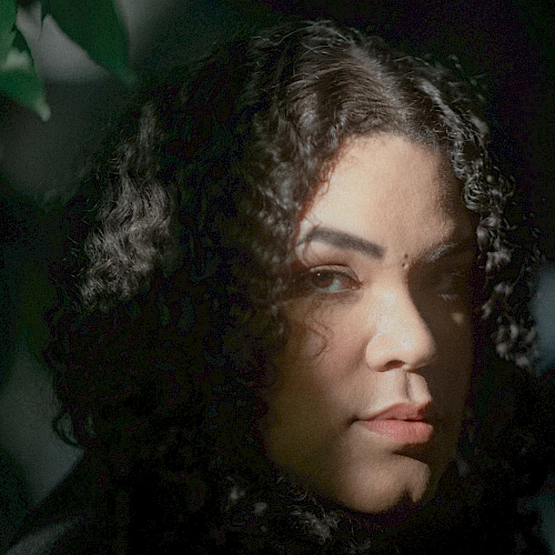 A person with curly hair is partially illuminated by light filtering through leaves, creating a dramatic, shadowed effect on their face.