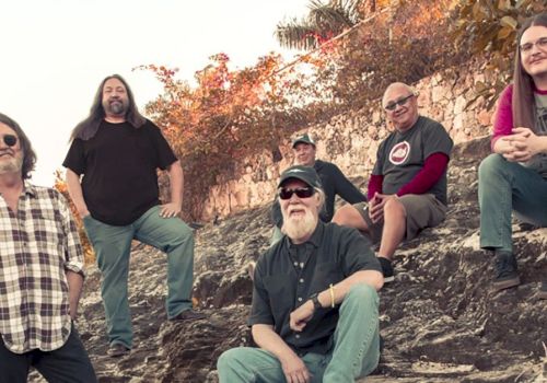 A group of six adults sitting and standing outdoors on a rocky hill with trees and a stone wall in the background.