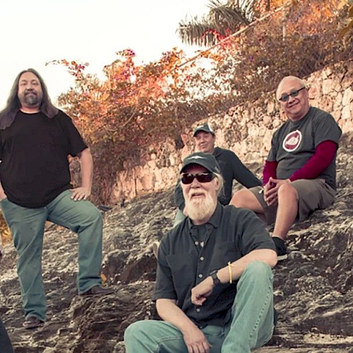 A group of six adults sitting and standing outdoors on a rocky hill with trees and a stone wall in the background.