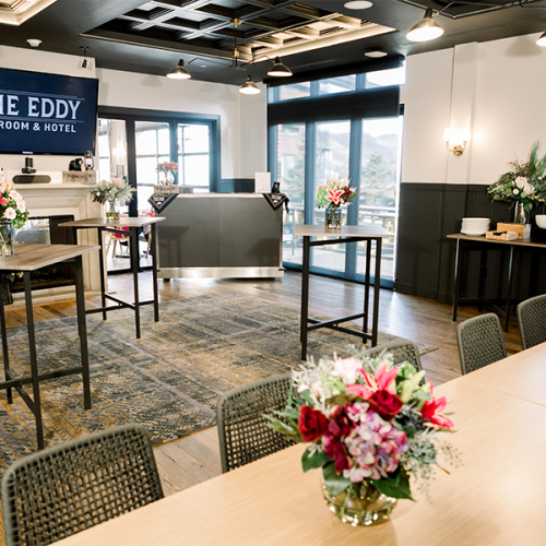 The image displays a chic taproom with floral arrangements, tables, and a sign for "The Eddy Taproom & Hotel" in a well-lit space.
