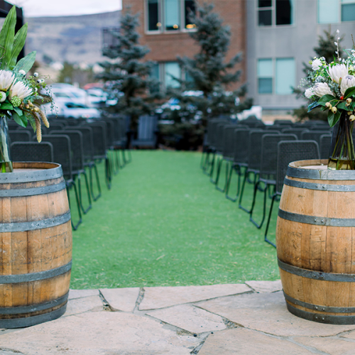 The image shows two wooden barrels with flower arrangements, set up in an outdoor venue with rows of chairs lined up.