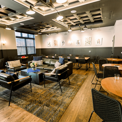 A modern lounge with seating, tables, artwork on the walls, and soft lighting, featuring wooden floors and decorative ceiling elements.