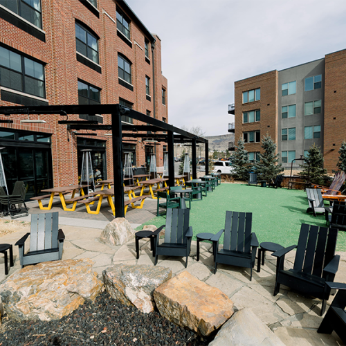 An outdoor seating area with black chairs, tables, a pergola, and artificial turf between brick buildings and rocks in the foreground.