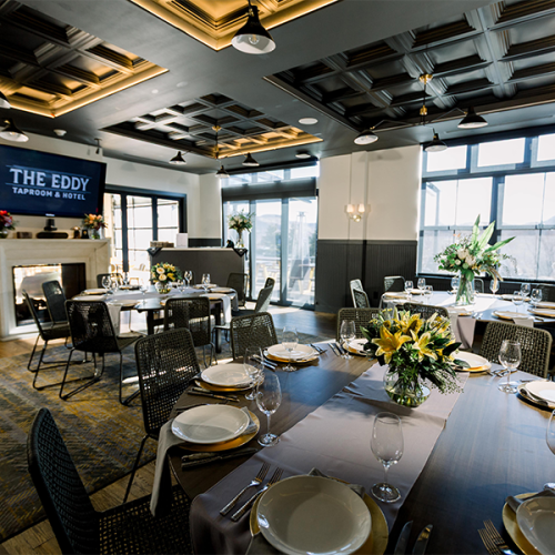 A set dining space with elegant tables, floral arrangements, and a sign reading "The Eddy," featuring large windows and stylish decor.