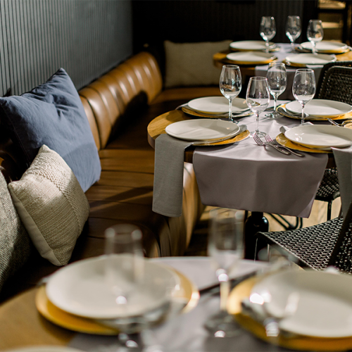 The image shows a cozy restaurant setting with tables set with plates, glasses, and cutlery, surrounded by cushioned chairs and sofas.