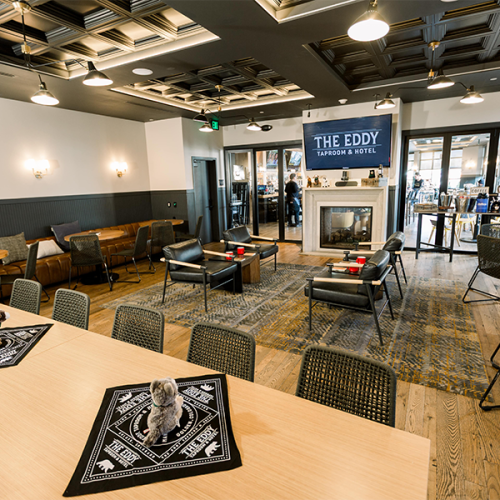 The image shows a modern lounge area with tables, chairs, and a fireplace at The Eddy Taproom & Hotel, creating a cozy ambiance.