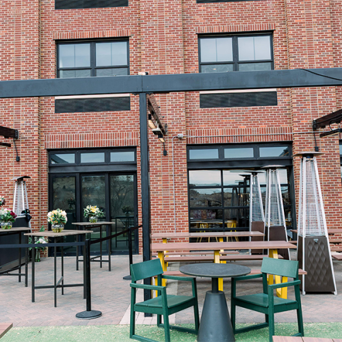 Outdoor patio with tables, chairs, and heaters in front of a brick building with large windows and black accents.