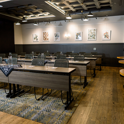 The image shows a conference room with rows of tables and chairs, decorated walls, and well-lit with wall-mounted lights and ceiling fixtures.