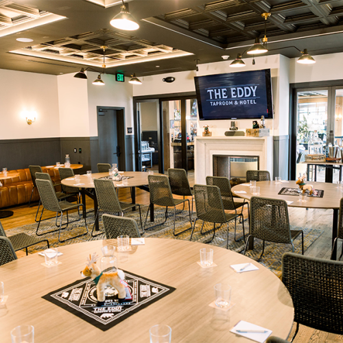 The image shows a dining area with round tables, chairs, and a sign for "The Eddy Taproom & Hotel" above a fireplace.