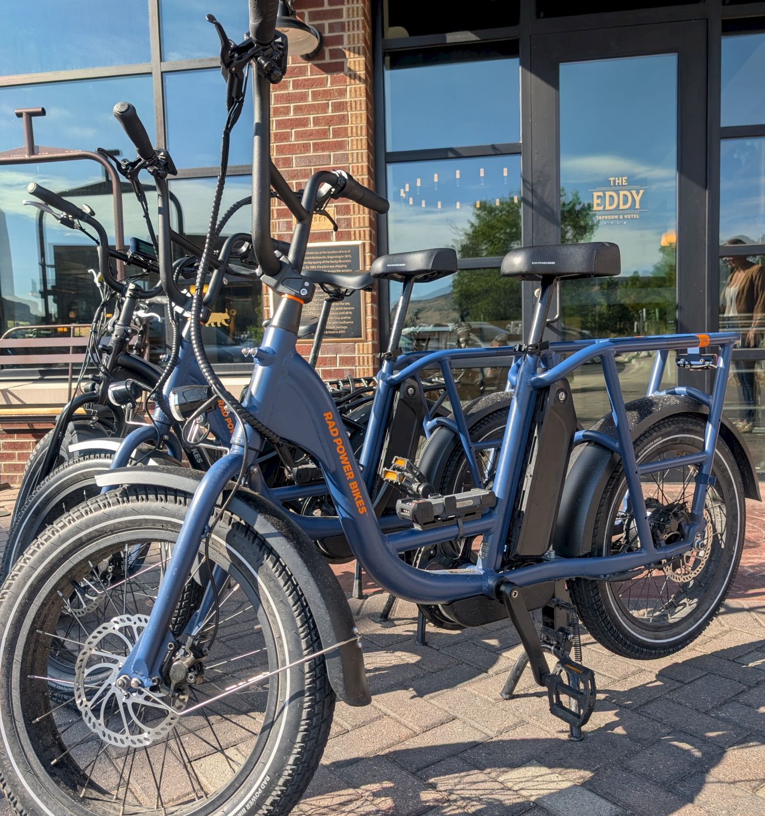 The image shows blue electric cargo bikes parked on a brick sidewalk near a building with large windows.