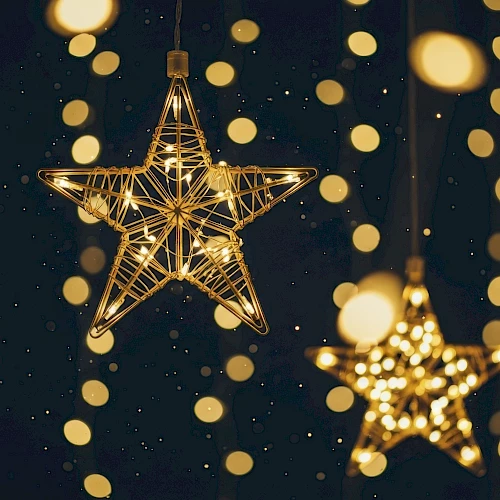 The image shows glowing star-shaped decorations hanging among blurred, circular lights on a dark background, creating a festive atmosphere.