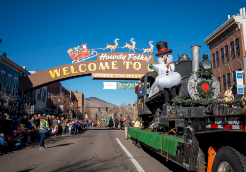 A festive parade with a decorated float under a "Howdy Folks! Welcome" sign, featuring a snowman and reindeer above the street.