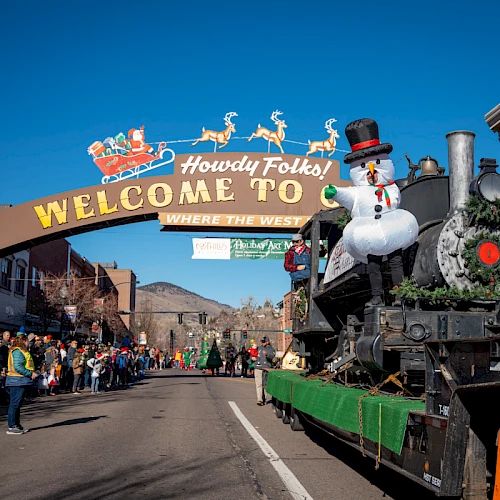 A festive parade with a decorated float under a "Howdy Folks! Welcome" sign, featuring a snowman and reindeer above the street.