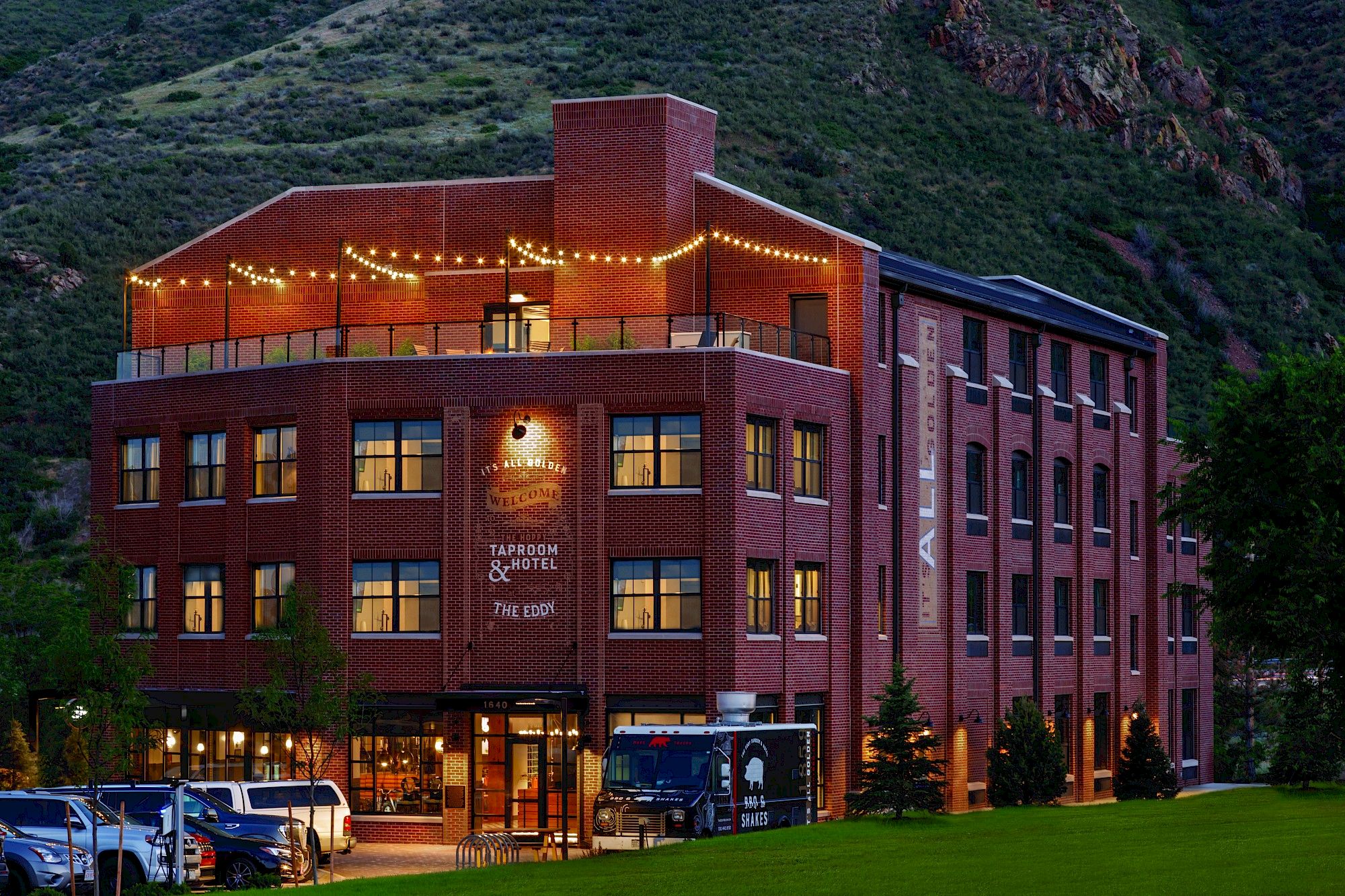 A brick building with string lights on the roof deck, identified as a tavern and hotel, surrounded by greenery and hills.