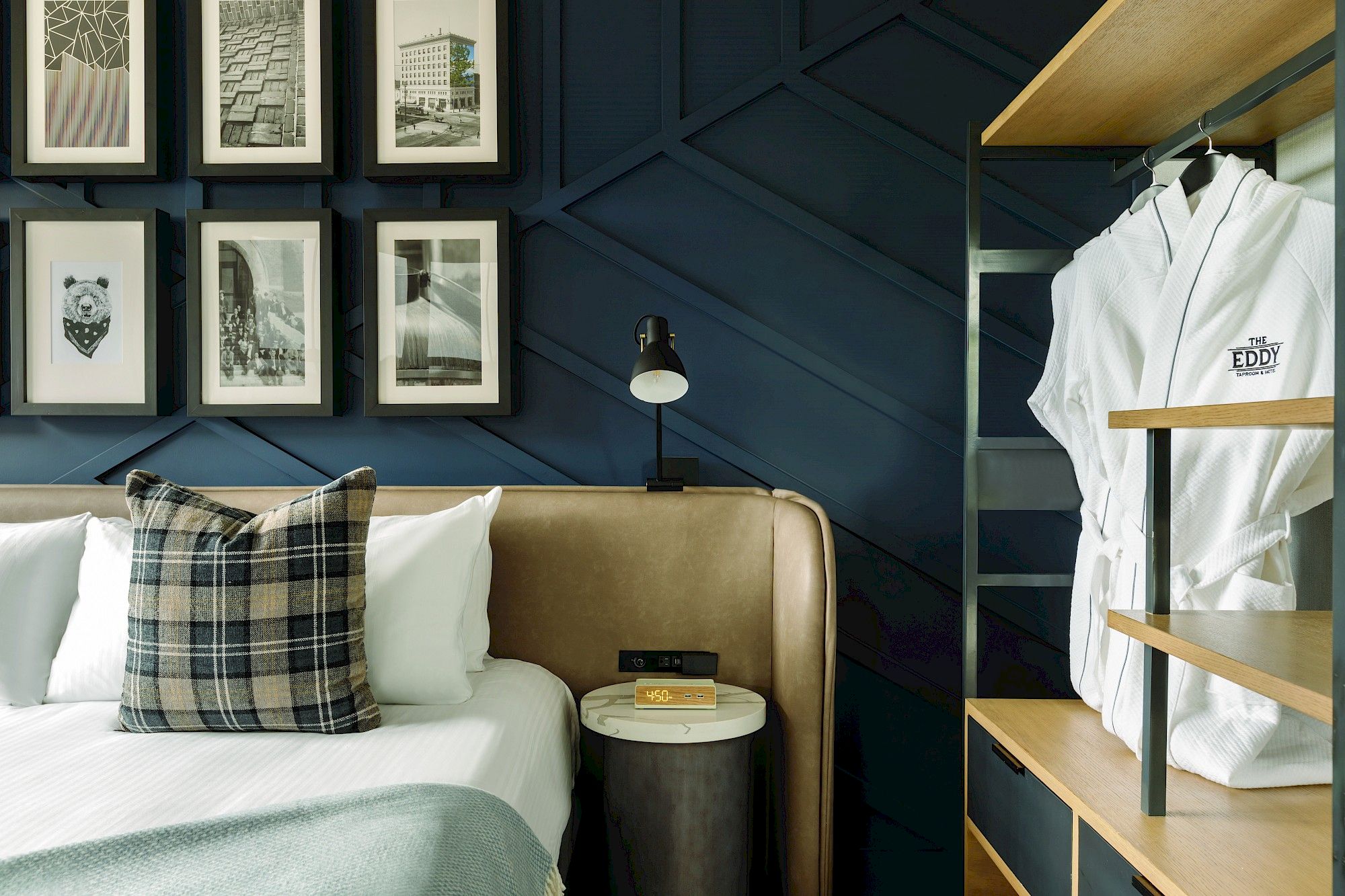 A stylish bedroom with framed artwork, a beige headboard, plaid pillows, a small nightstand with a book, and white robes hanging in an open closet.
