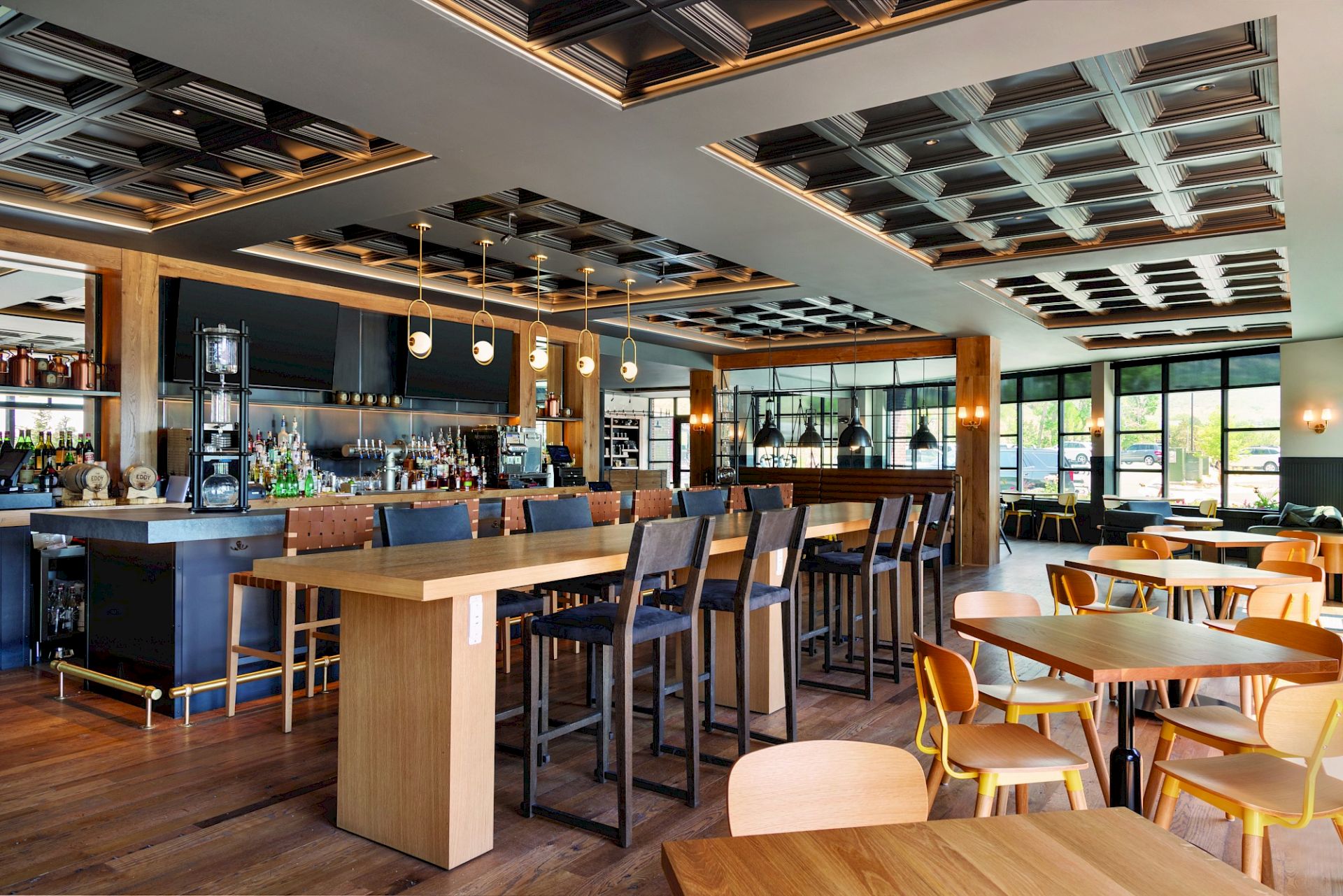 The image shows a modern, stylish restaurant interior with wooden tables, chairs, a long bar counter, ceiling lights, and large windows.