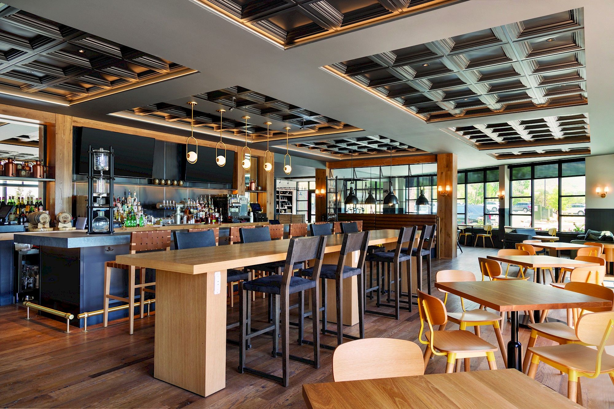 The image shows a modern, stylish restaurant interior with wooden tables, chairs, a long bar counter, ceiling lights, and large windows.