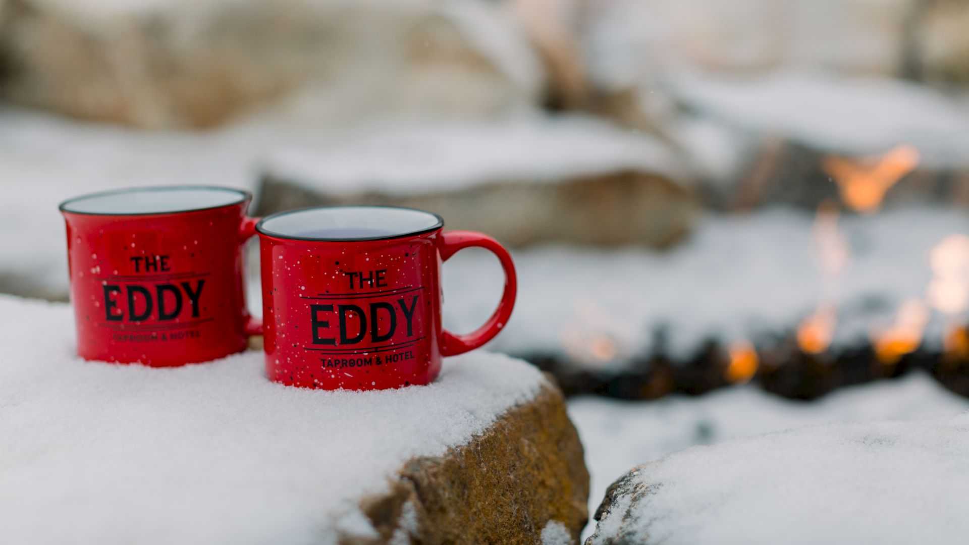 Red mugs labeled "The Eddy" sit on snow-covered rocks near a fire pit.