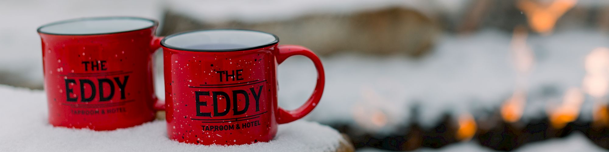 Red mugs labeled "The Eddy" sit on snow-covered rocks near a fire pit.