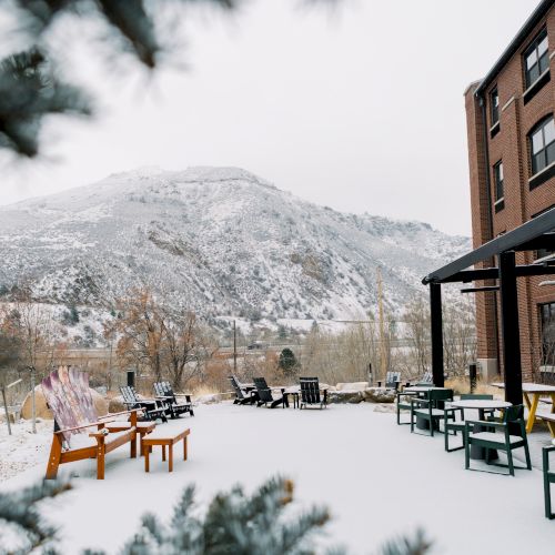 A snowy outdoor patio with tables and chairs, next to a brick building and a mountain in the background, framed by pine branches.