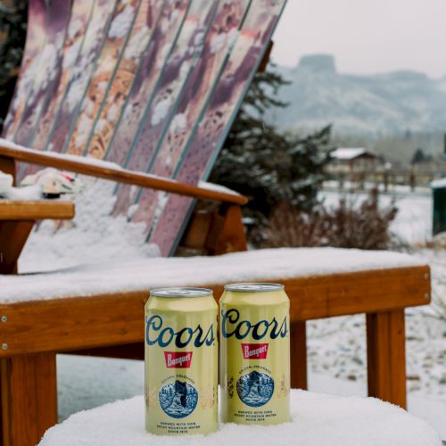 The image shows two Coors beer cans on a snowy table, with a bench made of skis in the background and a snowy landscape.