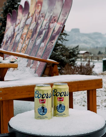 Two Coors beer cans sit on a snowy table with a wooden bench made of skis in the background.