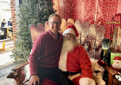 A person sits on Santa's lap in a festive setting with skis and red decorations in the background.