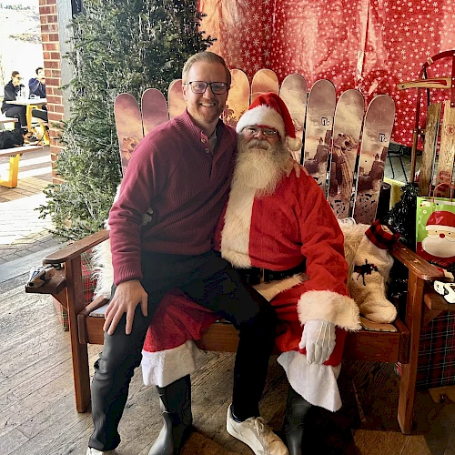 A person sits on Santa's lap in a festive setting with skis and red decorations in the background.