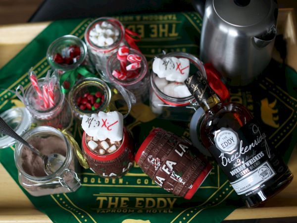 Assorted bar snacks and drinks on a wooden tray: liqueurs, jelly beans, marshmallows, candy, a bottle of beer, and a metal kettle, all on a green cloth.