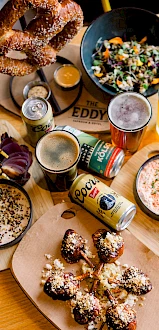 A spread of beer, pretzels, spicy fried bites, dips, salad, and a colorful assortment of dishes on a wooden table, perfect for a casual feast.