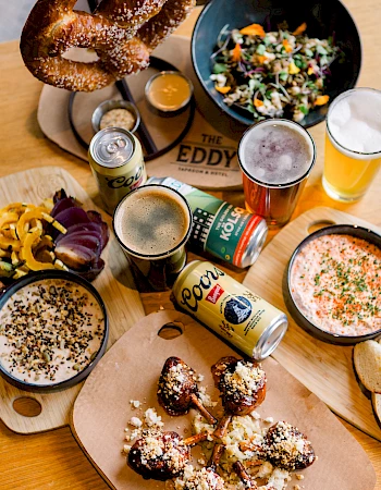 A spread of beer, pretzels, spicy fried bites, dips, salad, and a colorful assortment of dishes on a wooden table, perfect for a casual feast.