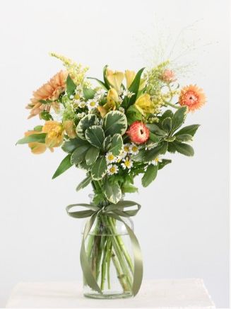 A bright bouquet of yellow, orange, and white flowers in a glass vase with greenery and a ribbon, set against a white background.