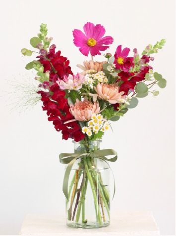 A bright bouquet with pink, red, and cream flowers in a clear jar, tied with a green ribbon.