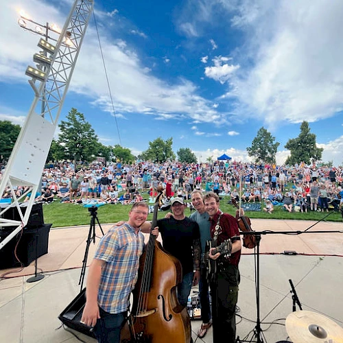 A band poses with instruments on an outdoor stage in front of a large cheering audience under a blue sky with scattered clouds.