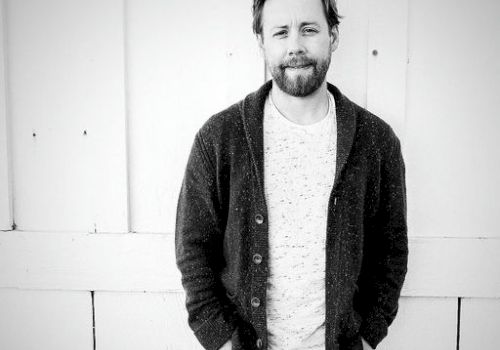 A bearded man in a cardigan and white shirt stands casually against a white paneled wall, hands in pockets, outdoors.