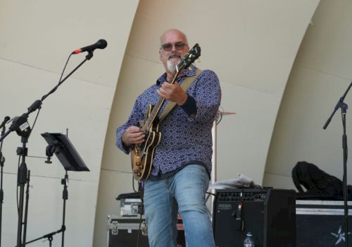 A bearded man wearing a patterned shirt playing an electric guitar on a stage with microphones and equipment, performing live.