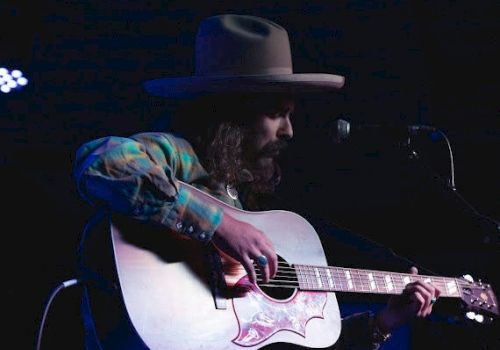 A bearded musician wearing a wide-brim hat plays an acoustic guitar on stage.